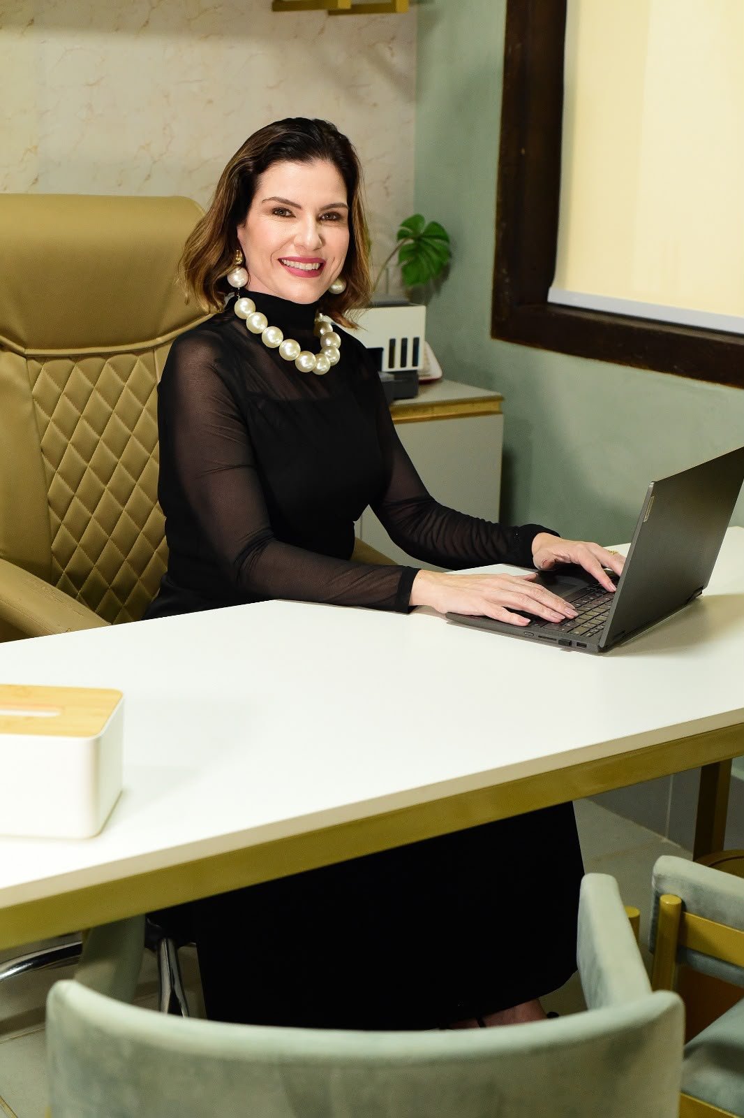 Dra. Ana Carla Prado sentada à mesa do consultório, junto ao notebook